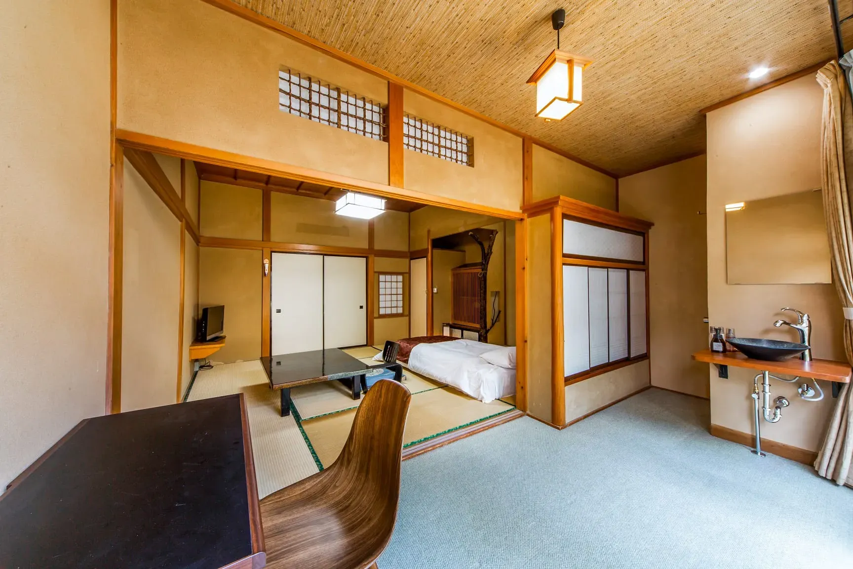 A hotel guest room that fuses traditional Japanese design with modern amenities. A raised platform with tatami mat flooring holds a low table and a bed with white linens. The lower, carpeted area in the foreground contains a modern wooden desk and chair, and a vanity with a black vessel sink.
