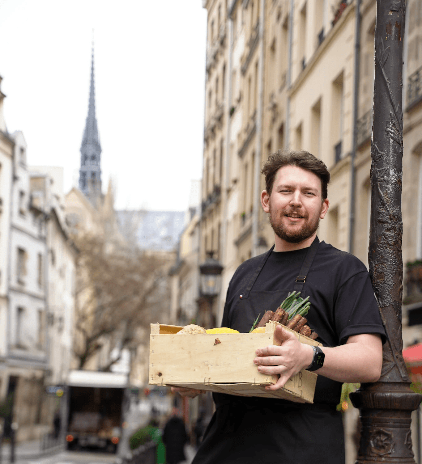 Chef Wilfried Romain devant le restaurant LAVA à Paris, tenant une caisse de produits frais dans le Quartier Latin, illustrant une cuisine de saison et l’approvisionnement en produits de qualité.