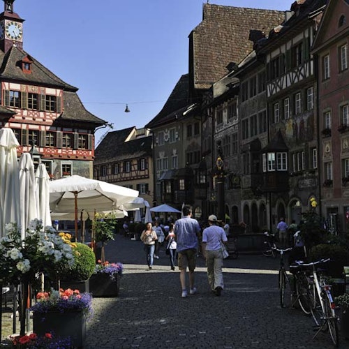 People walking on a cobblestone street in a European town with historic buildings, outdoor cafe seating, and bicycles.