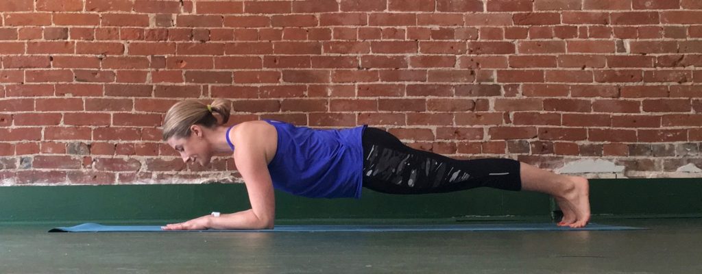 Heather Hausman in athletic wear is performing a forearm plank on a yoga mat indoors, with a background of exposed brick walls.