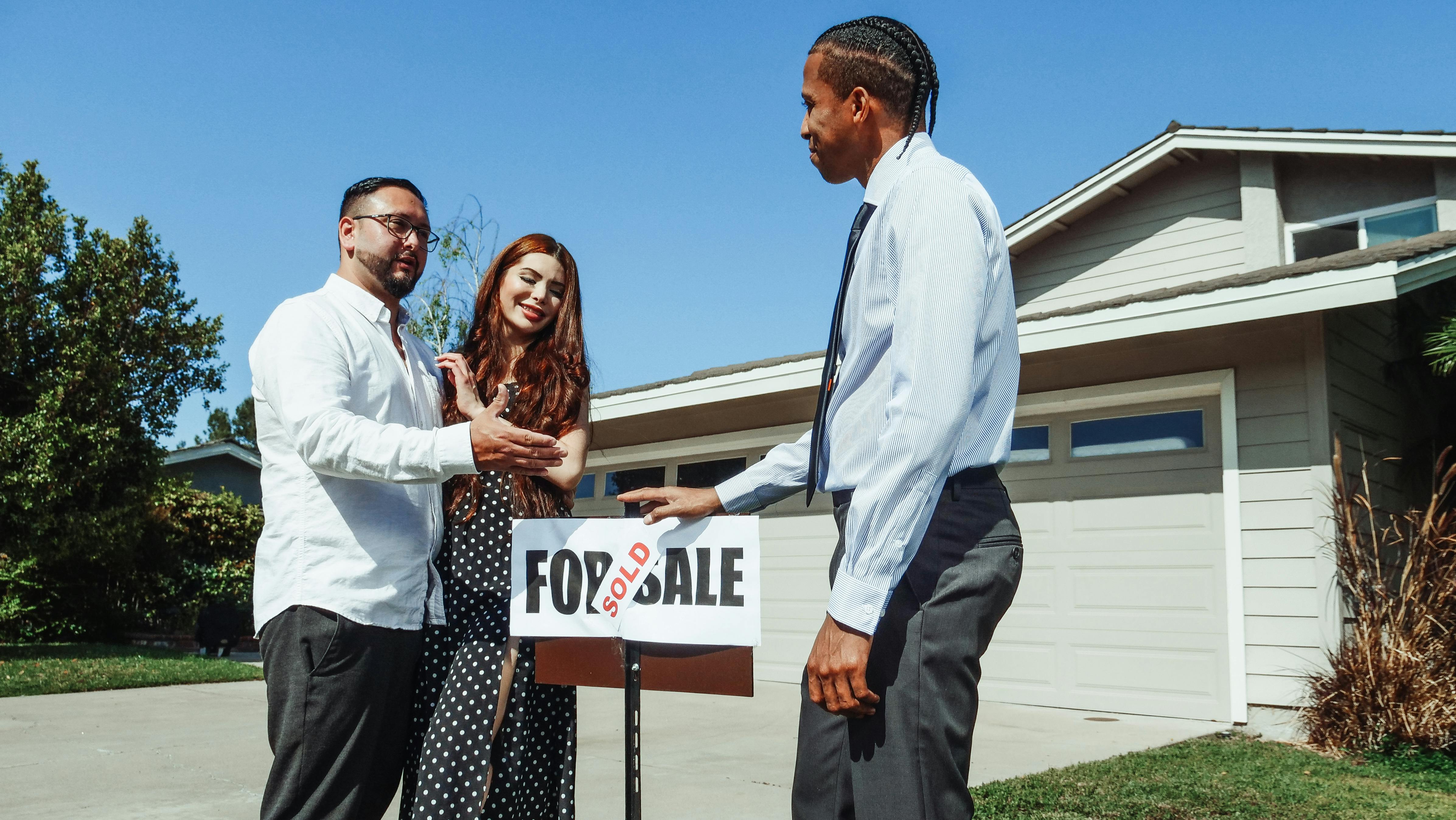 A couple stands in front of a house, shaking hands with a real estate agent next to a "Sold" sign.