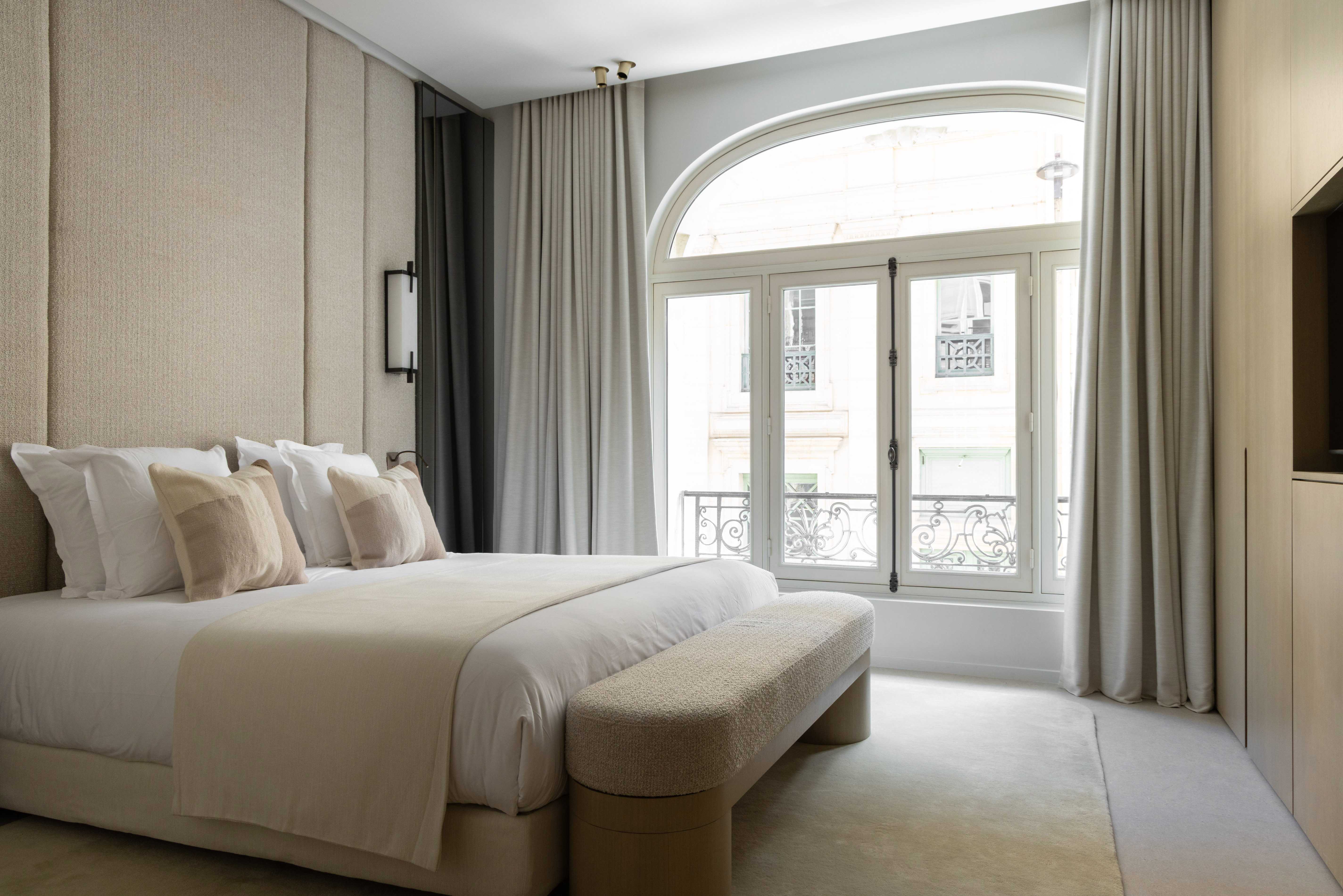 Chambre principale avec grand lit, tete de lit haute en tissu beige, banc en bout de lit et fenetre cintre sur balcon