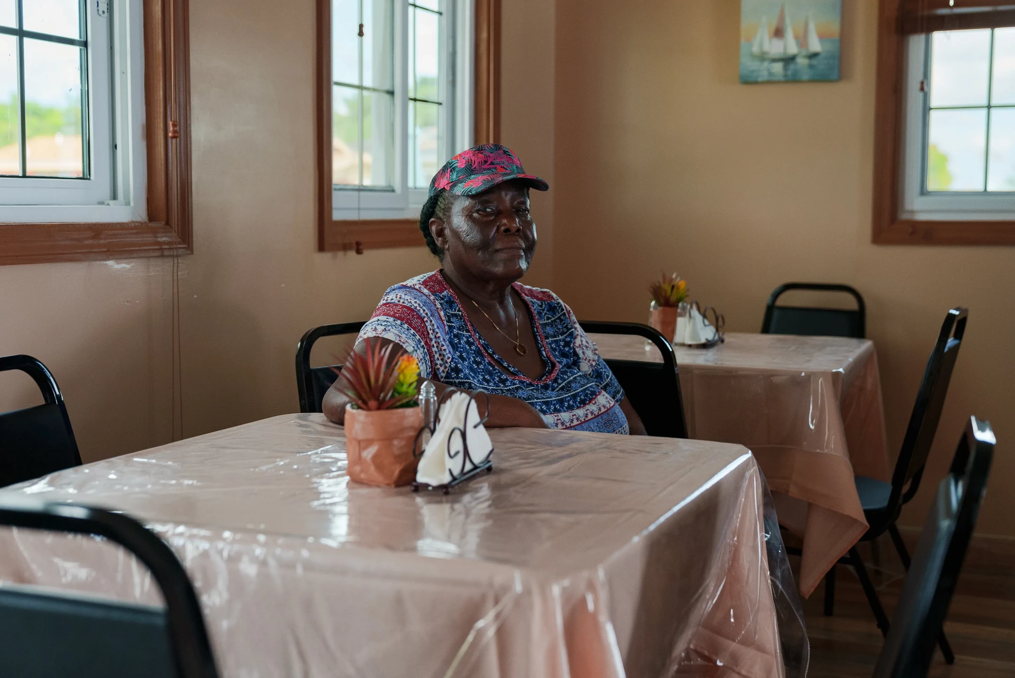 A person wearing a colorful shirt and cap sits at a table covered with a plastic tablecloth in a cozy, warmly lit room. The room has several tables with chairs and nautical-themed decor on the walls.