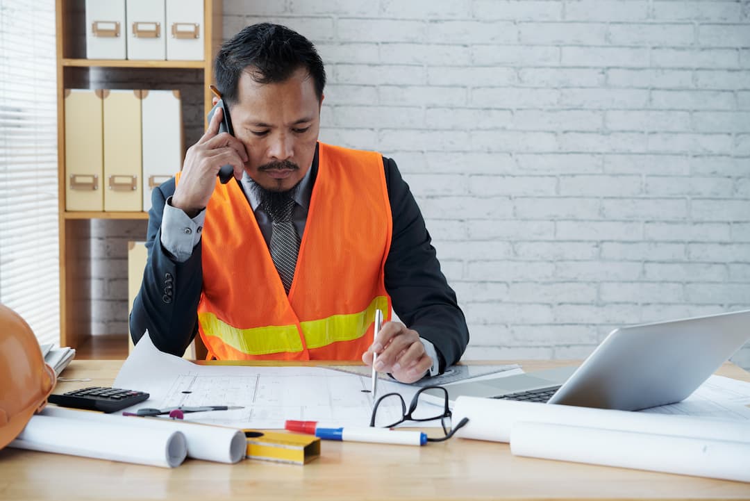 A construction professional wearing an orange safety vest sits at a desk while talking on a phone. There is a laptop, glasses, and rolled blueprints.