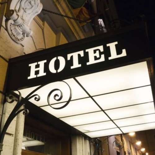Insegna luminosa dell'hotel con staffe di metallo decorative, montata sopra un ingresso. Dettaglio dell'edificio visibile sullo sfondo.