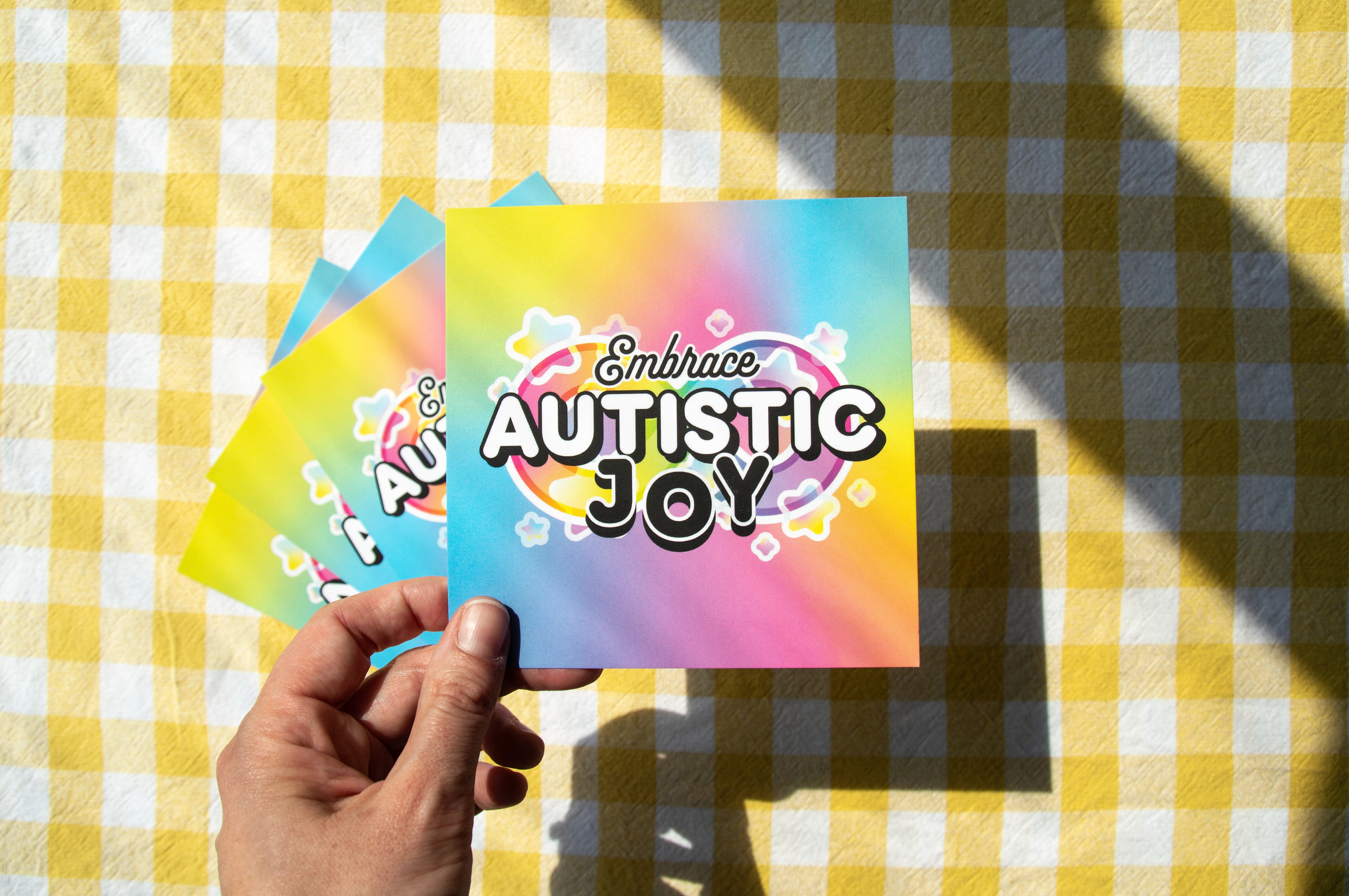 A hand holding a small square print. It is coloured in rainbow pastels and states "Embrace Autistic Joy". Behind the text is an infinity symbol surrounded by stars.