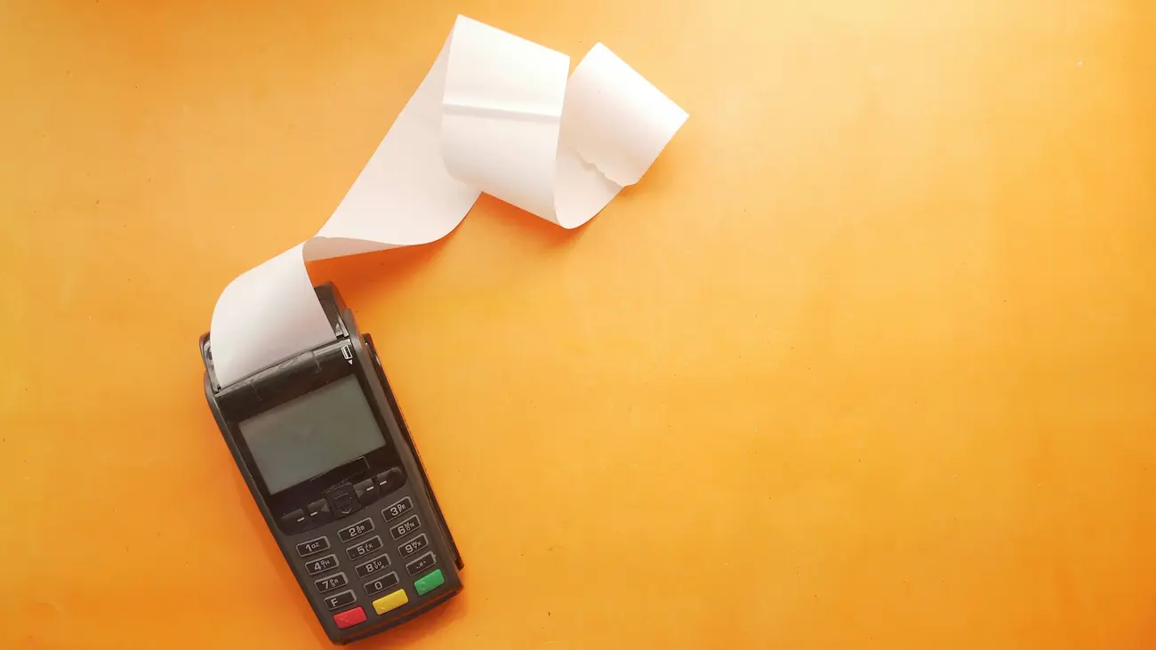 Card payment terminal printing a long receipt on an orange background, illustrating the difference between a donation receipt and an invoice for sports club sponsorship.