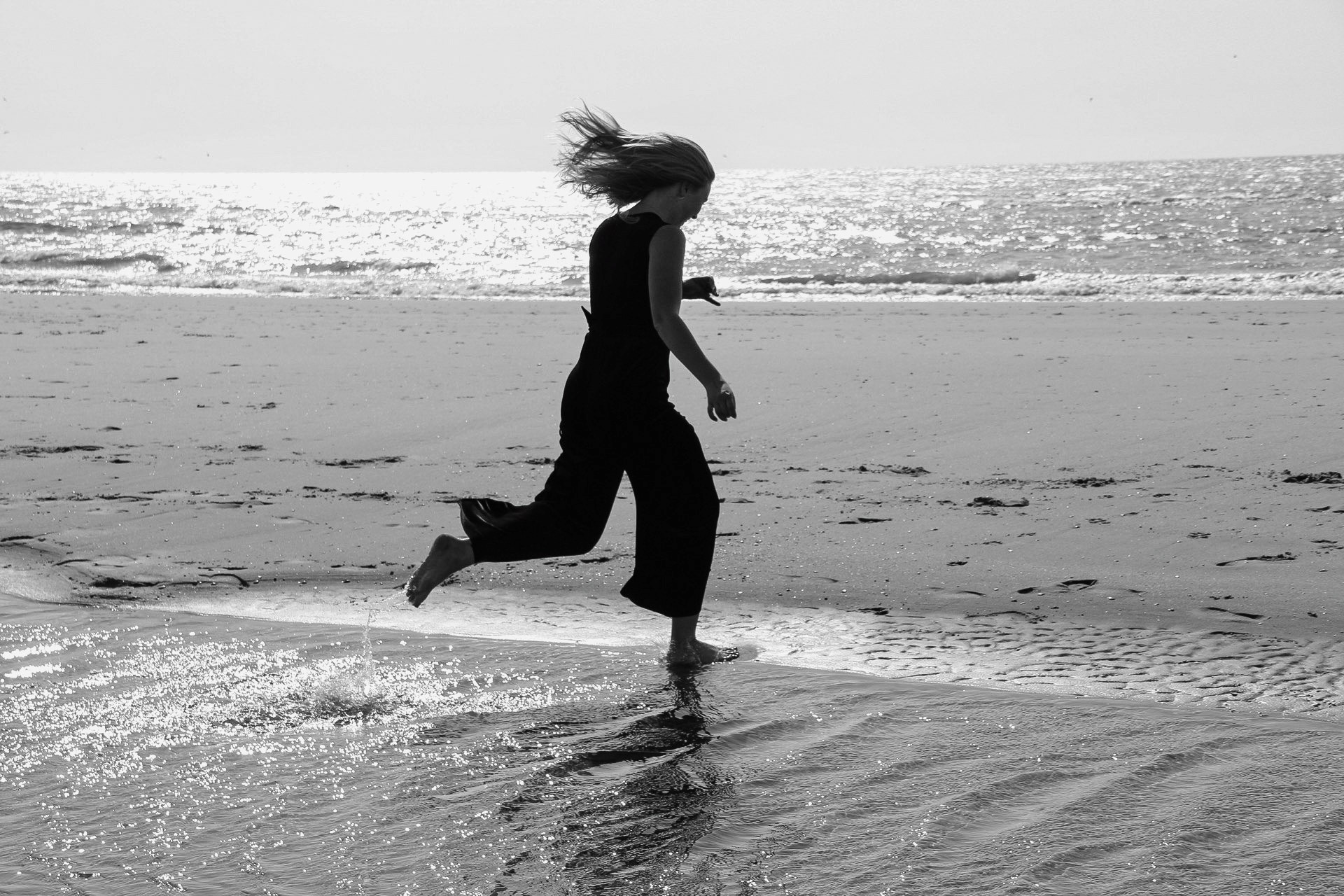 Dynamische foto van een vrouw die een sprongetje maakt over het water op het strand, met een focus op de beweging en de vreugde van het moment