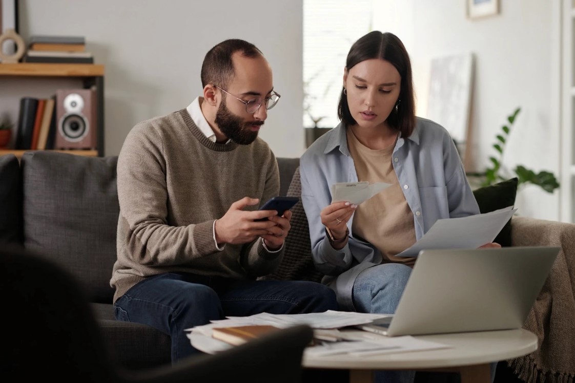 Couple looking at paperwork and receipts and using a cell phone while sitting on a couch.