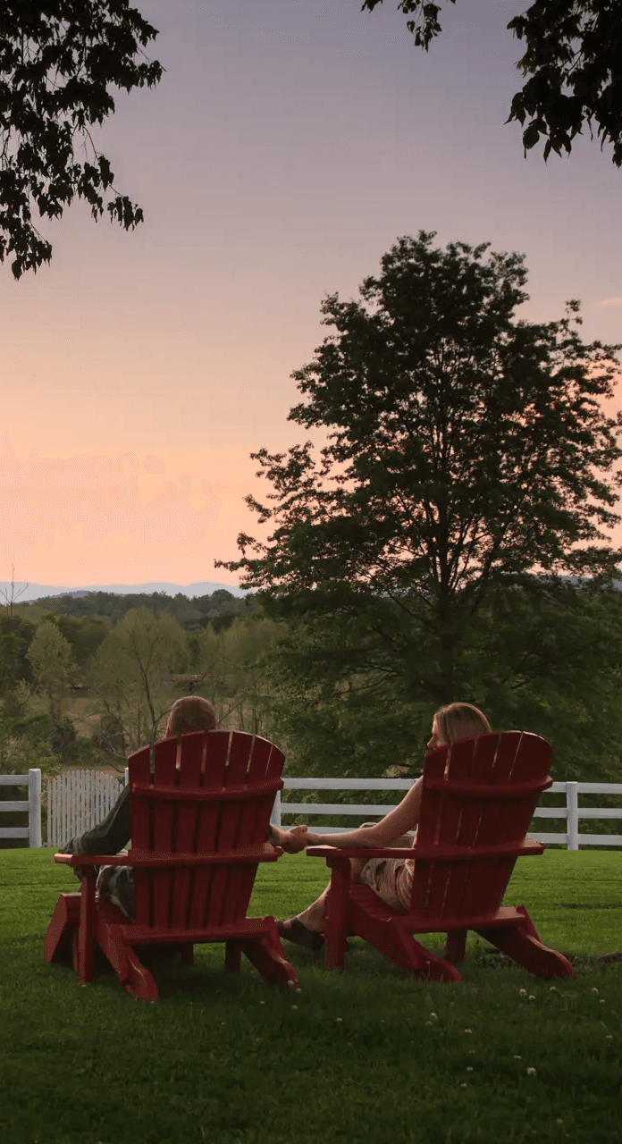 Two people sit in red Adirondack chairs on a lush green lawn, facing a serene sunset view with trees and rolling hills in the background at The Estate at Meander.