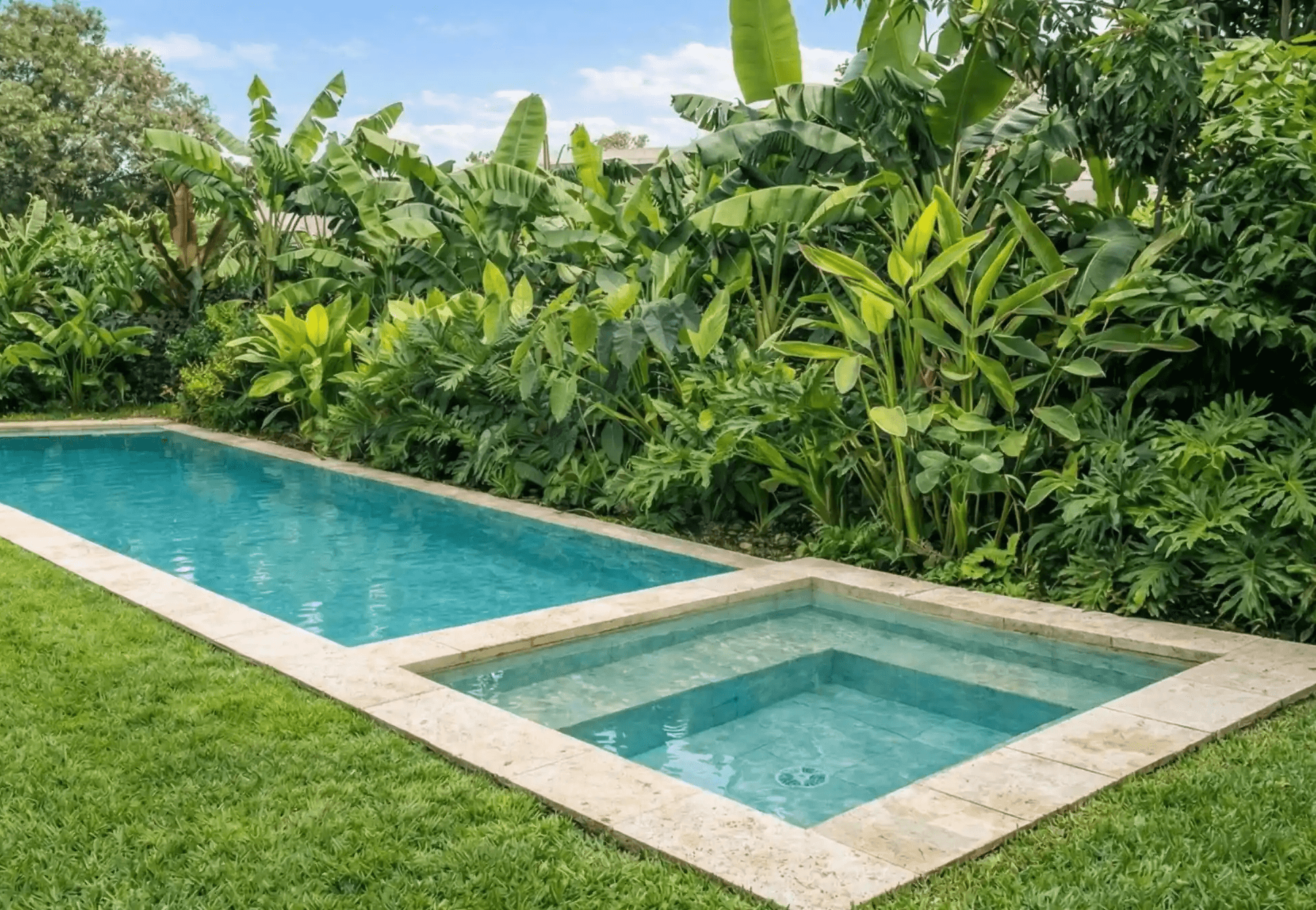 Piscina rectangular revestida en tonos claros con anexo para niños, rodeada de un exuberante jardín tropical con palmeras.