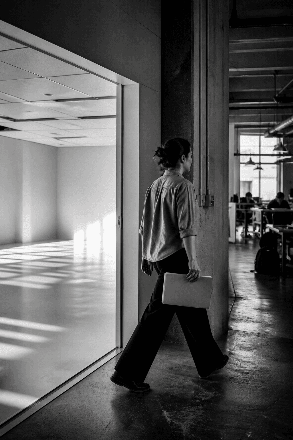 Black-and-white editorial photograph of an employee carrying a folder through a doorway from an empty pilot room into an active workplace.