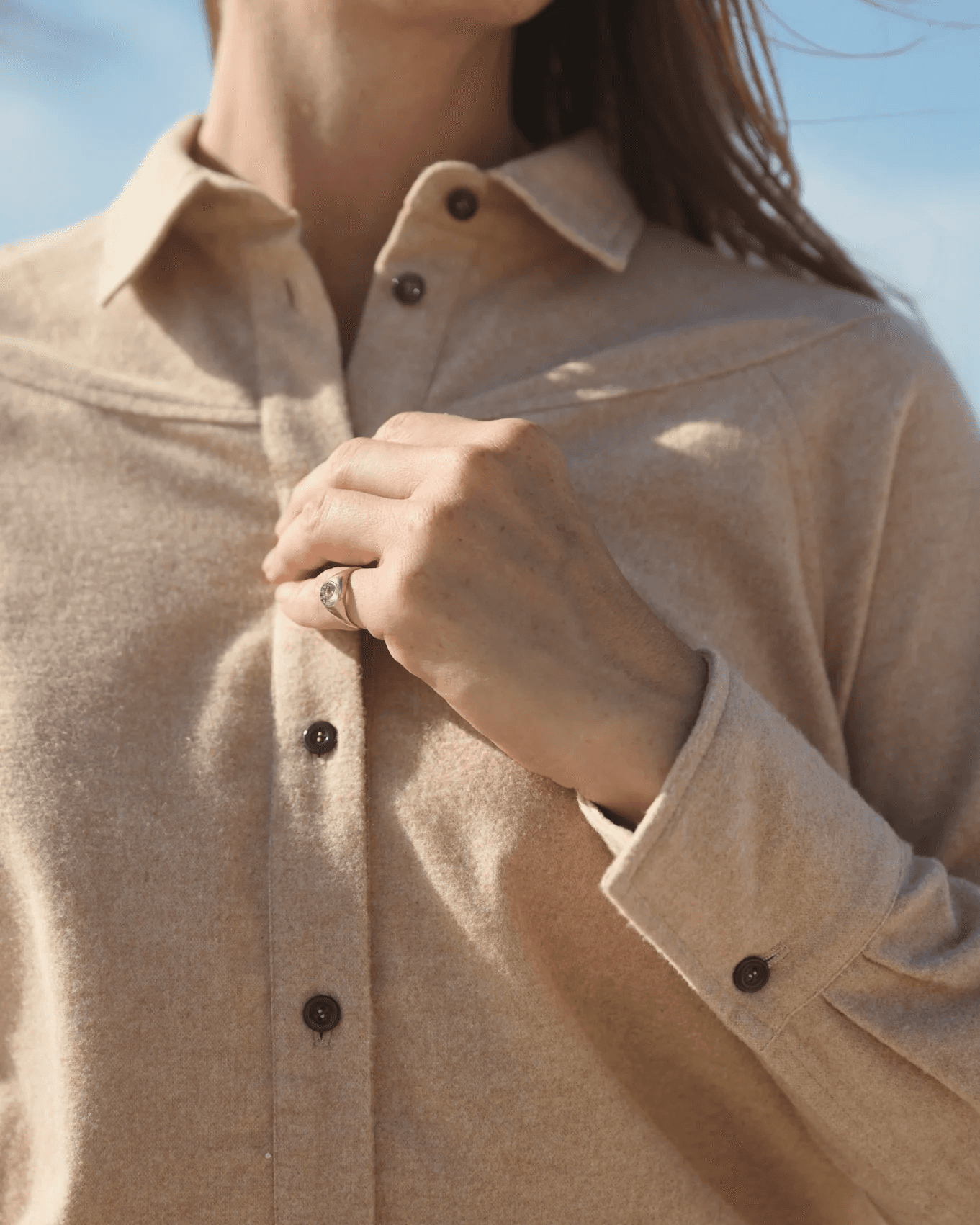 A person gently holds the front of a beige button-up wool shirt, showcasing a subtle design and a minimalist ring on their finger, against a clear blue sky background.