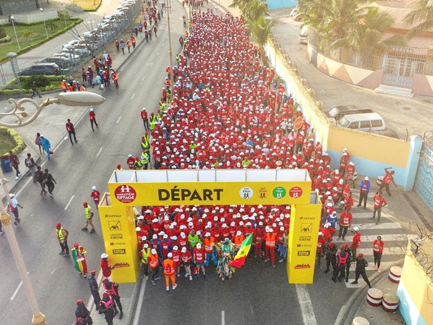 Vue aérienne d'une grande foule de coureurs sous l'arche de départ du marathon de Dakar.