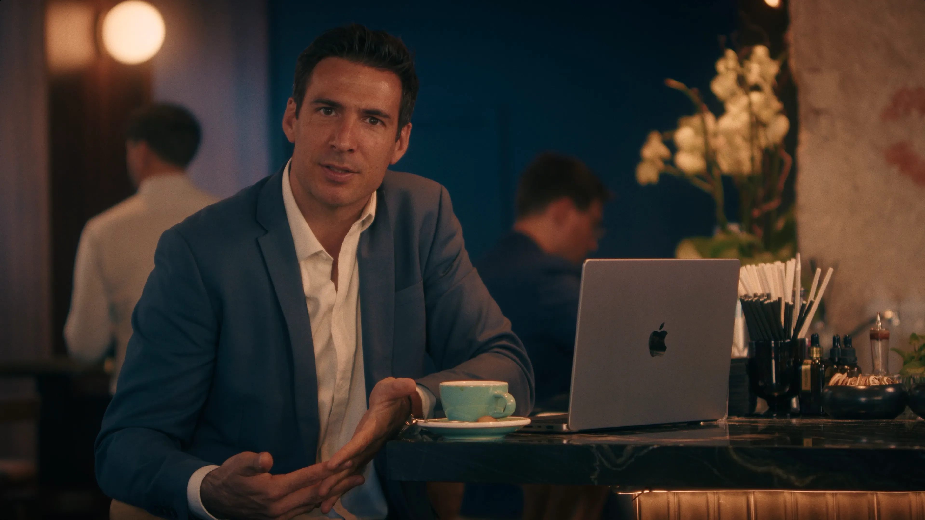 A man in a suit sits at a table with a laptop, looking engaged in conversation in a cozy cafe setting.