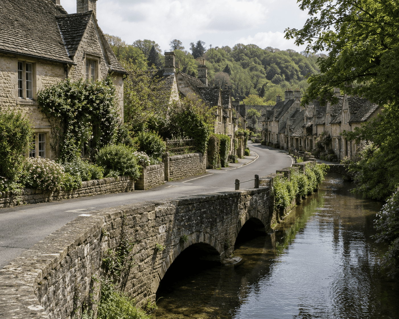 A scenic view of the picturesque village of Castle Combe in the Cotswolds, England. A historic arched stone bridge crosses the calm Bybrook river in the foreground, leading onto a narrow paved road lined with traditional honey-colored stone cottages, climbing greenery, and steep slate roofs against a backdrop of lush green wooded hills under a bright, cloud-filled sky.