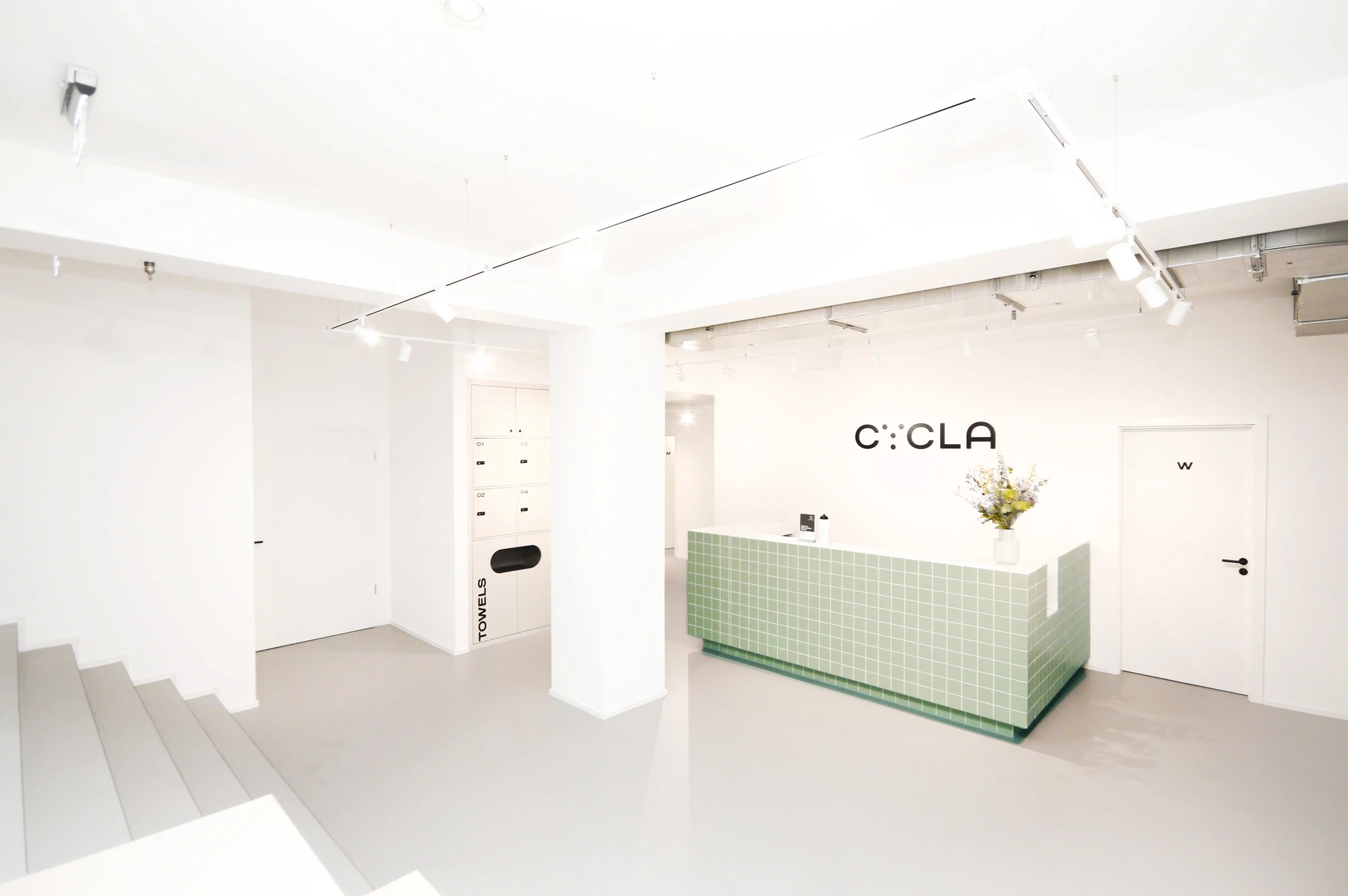 Minimalist Cycla reception area with mint-green tiled counter, white walls, locker zone labelled „Towels" and the Cycla logo on the back wall.