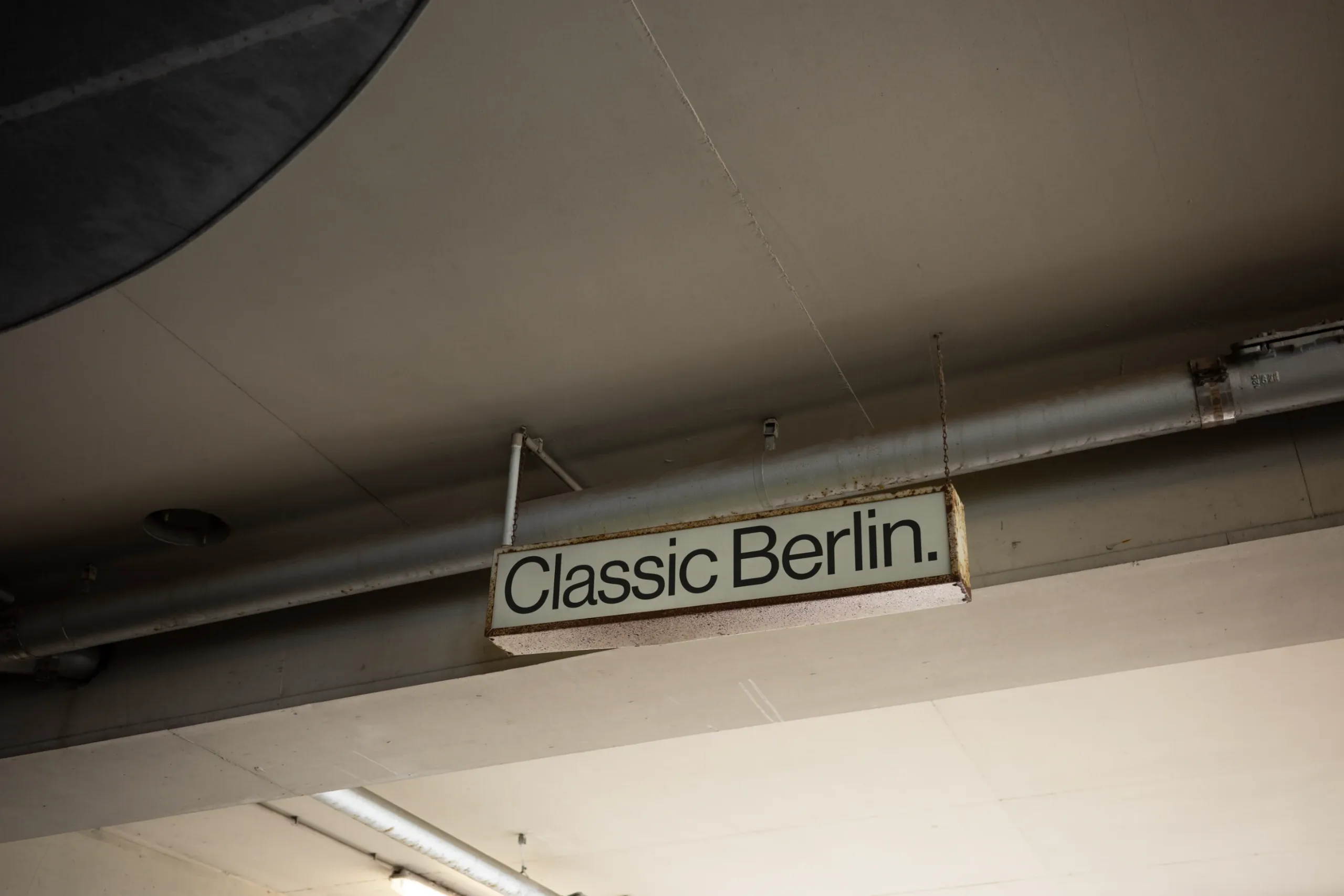 Ceiling-mounted directional sign in a minimalist garage interior. The sign features a grid design with black text, with visible aging and rust along the edges.