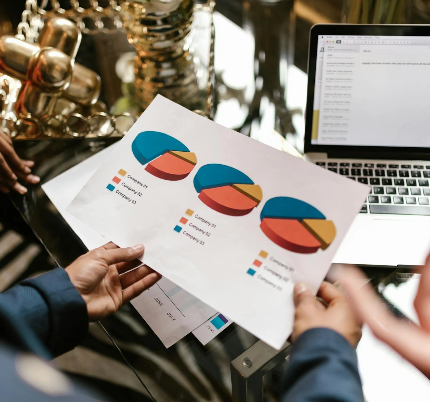 A hand holding a paper containing differnent pie charts in front of a laptop