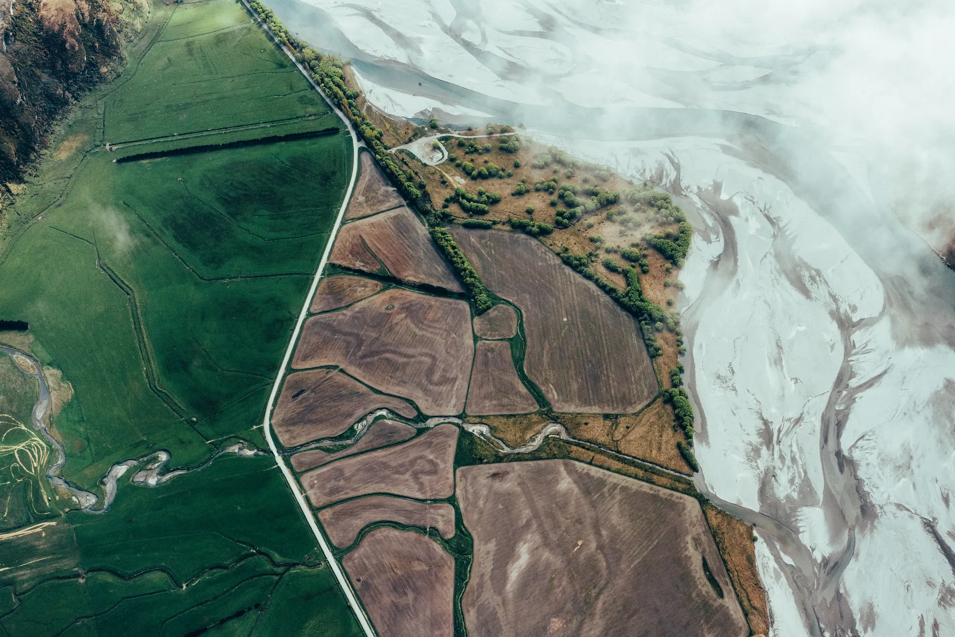 Aerial of green and ploughed farm paddocks meeting a pale braided riverbed