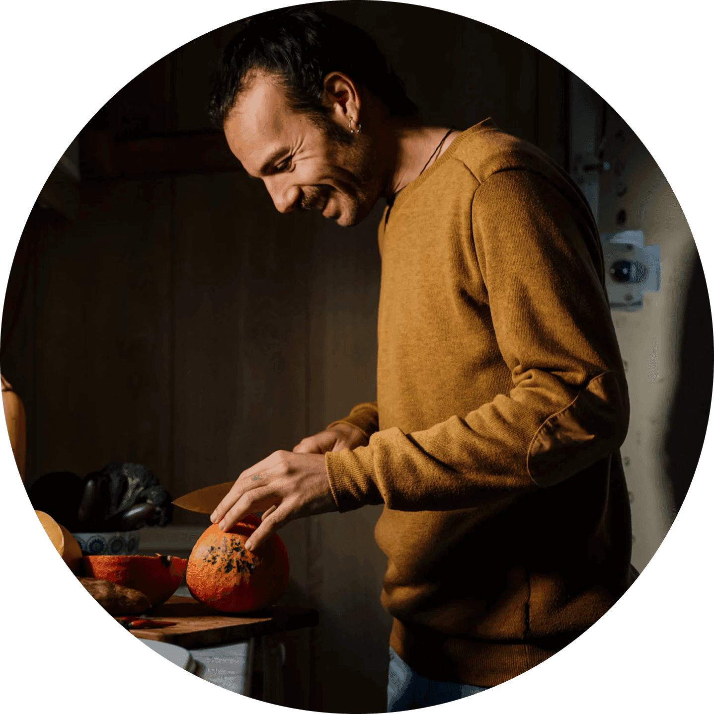 A man in a brown sweater prepares food in a warmly lit kitchen, smiling as he cooks.