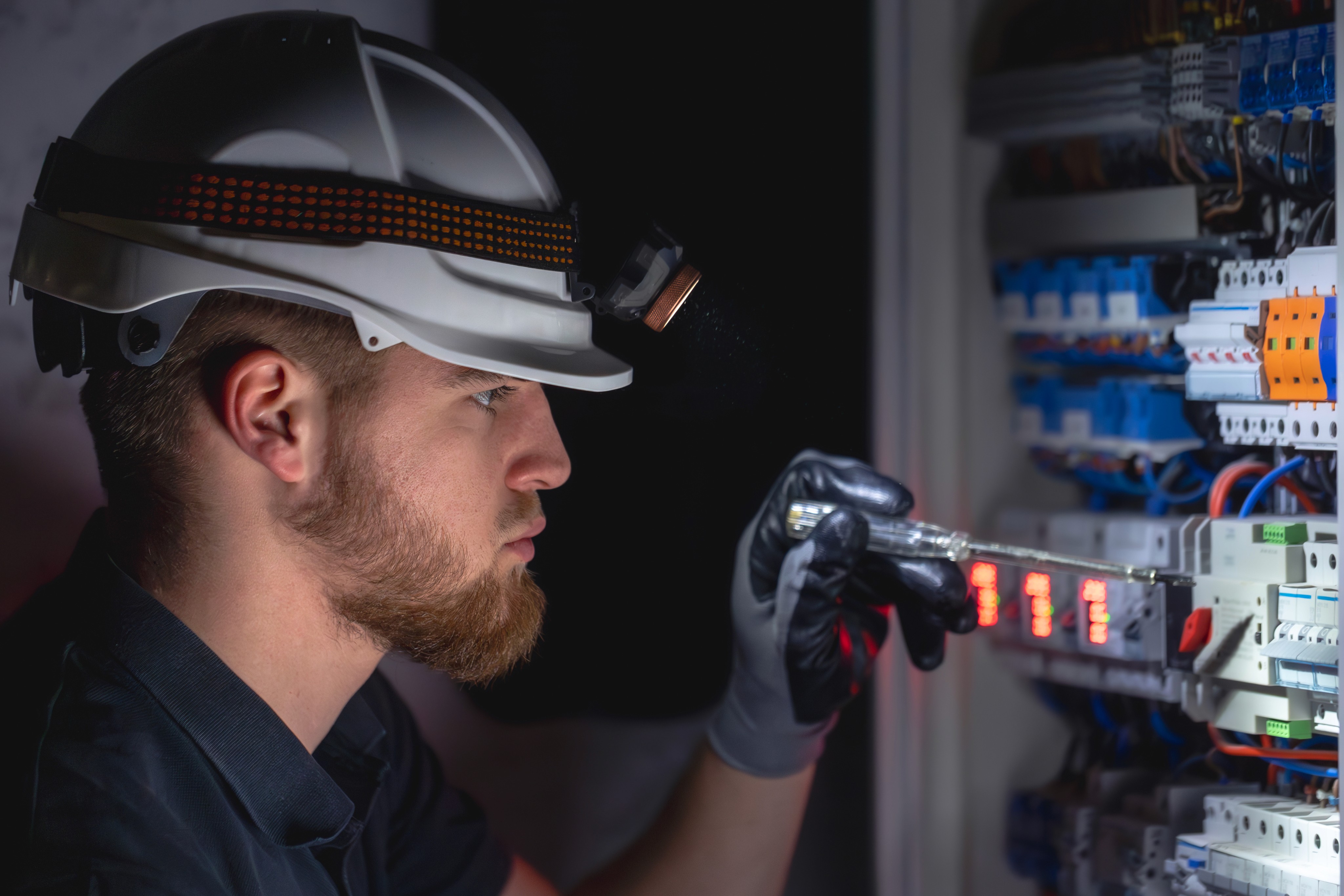 Electrician wearing a white hard hat with a headlamp inspecting an illuminated electrical panel at night