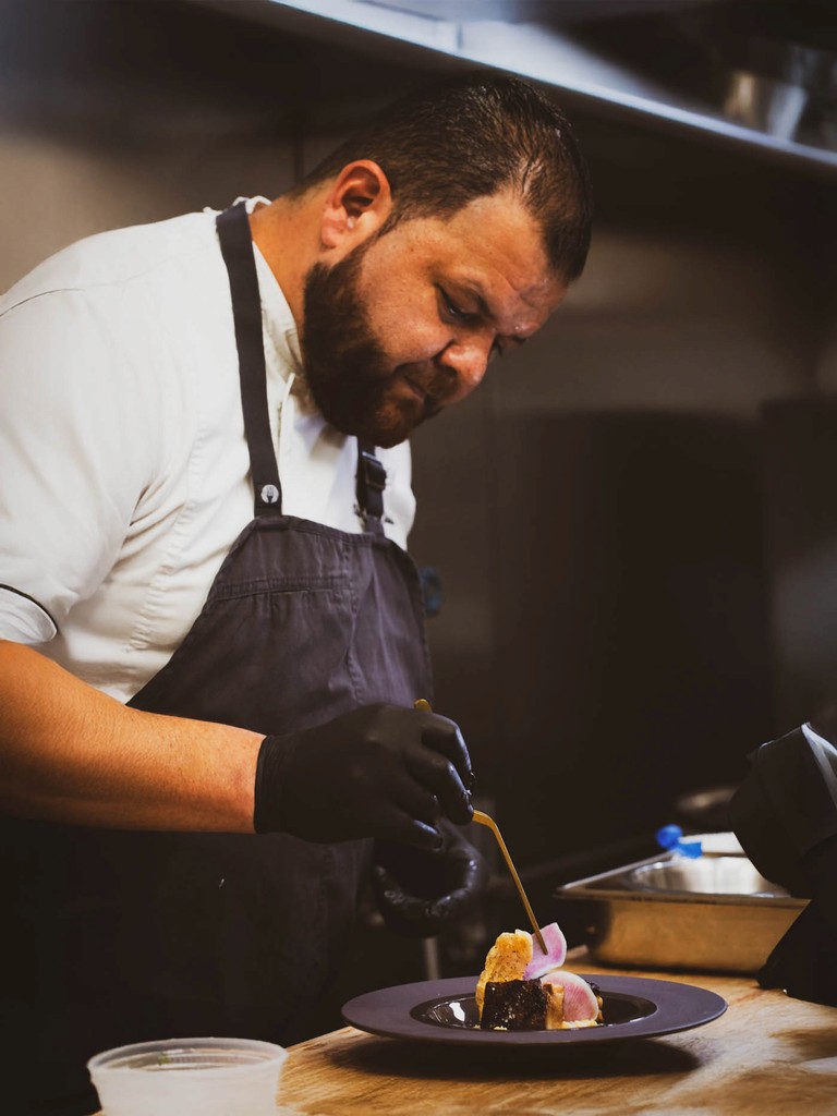 A male chef in a black apron carefully plates a dessert in a restaurant kitchen.