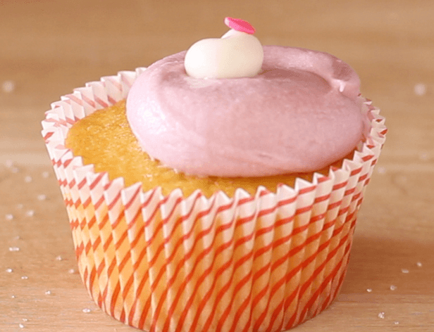 A single cupcake in a striped orange and white paper liner sits on a light wooden surface. The cupcake is topped with a swirled light pink frosting and a small white swirl on top. A small pink candy or sprinkle decoration is visible on the frosting. The liner has vertical red stripes.