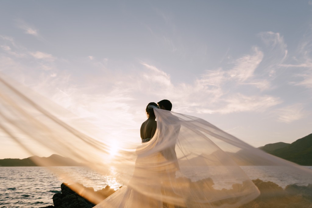 Bride and groom embracing at sunset with flowing veil over ocean