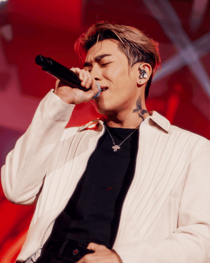 A male singer passionately performing on stage with a microphone, wearing a white jacket over a black shirt, with vibrant red lighting in the background.