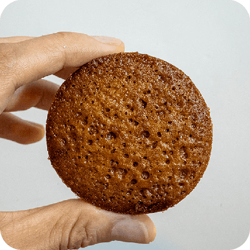 Primer plano de un brownie redondo sostenido con los dedos, resaltando su superficie caramelizada y textura esponjosa.