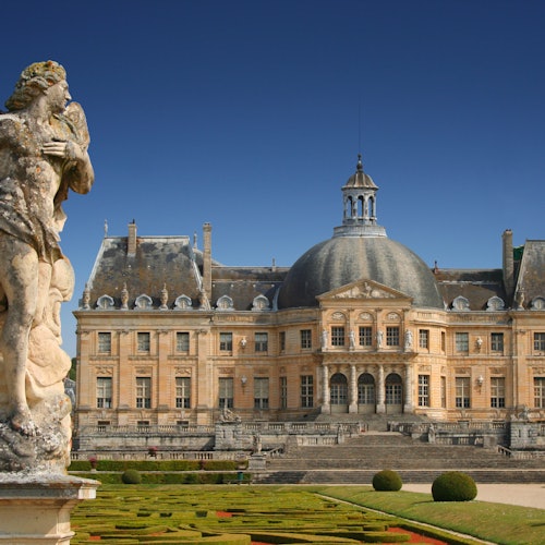 Статуя перед величественным историческим шато с большим куполом, ухоженными садами и чистым голубым небом.