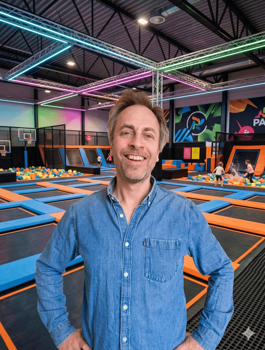 A young person with curly hair smiles at the camera while sitting in front of a laptop against a dark background.