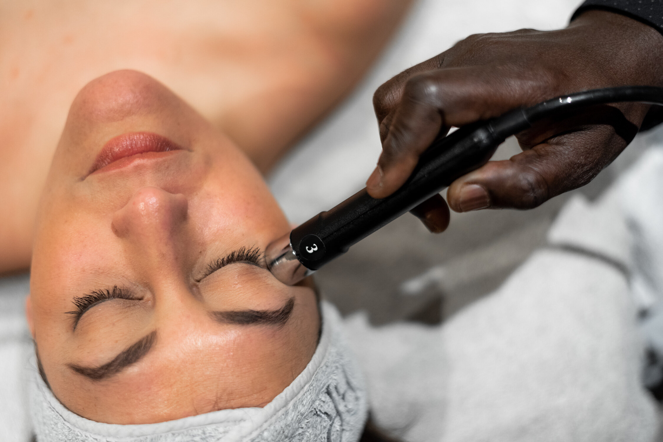 A person receives a facial treatment while another person uses an electronic device near their nose area.