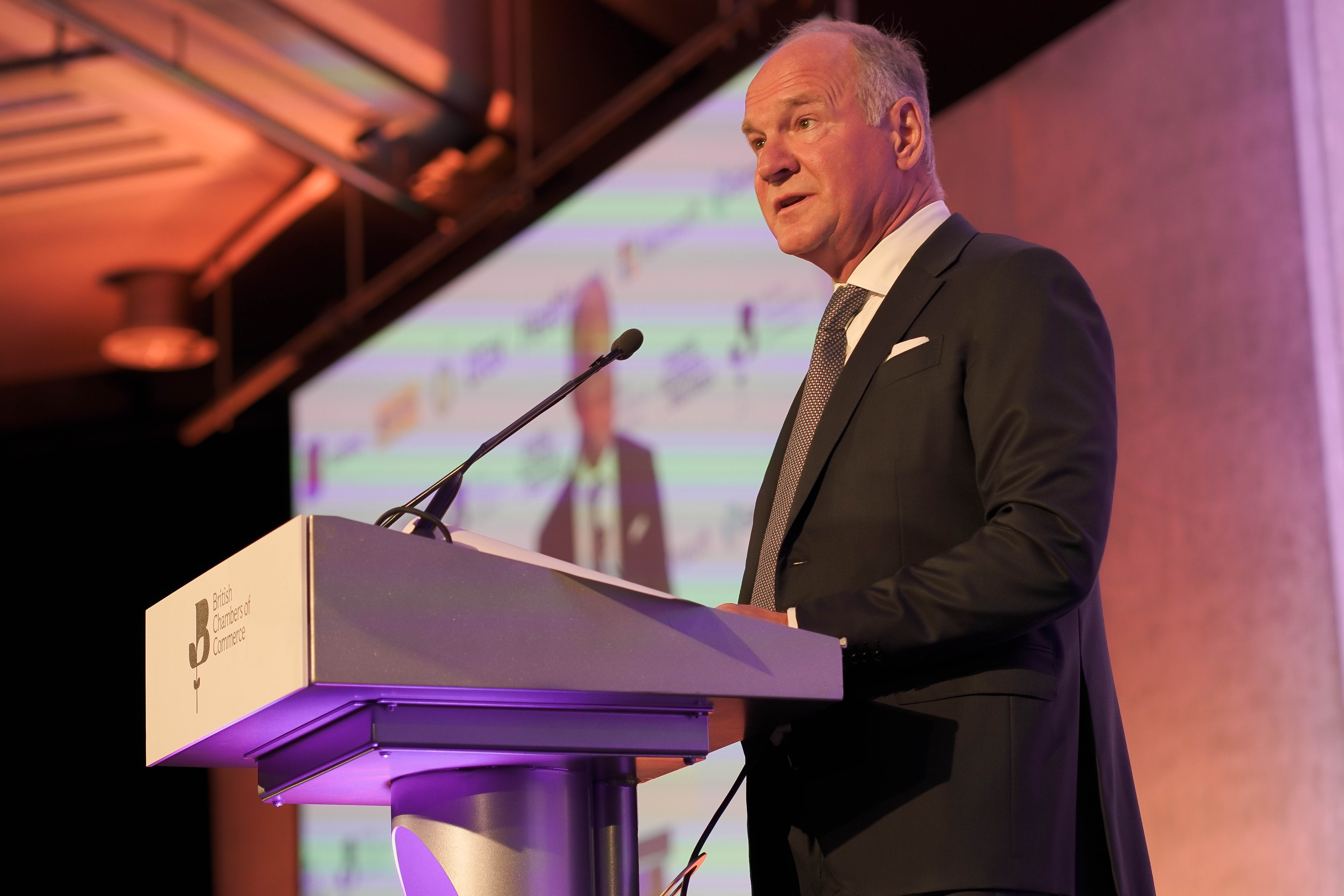 Heathrow CEO Thomas Woldbye, in a dark suit, delivers a speech from behind a podium with the British Chamber of Commerce logo on it. He is on a formally lit stage with a large screen behind him.