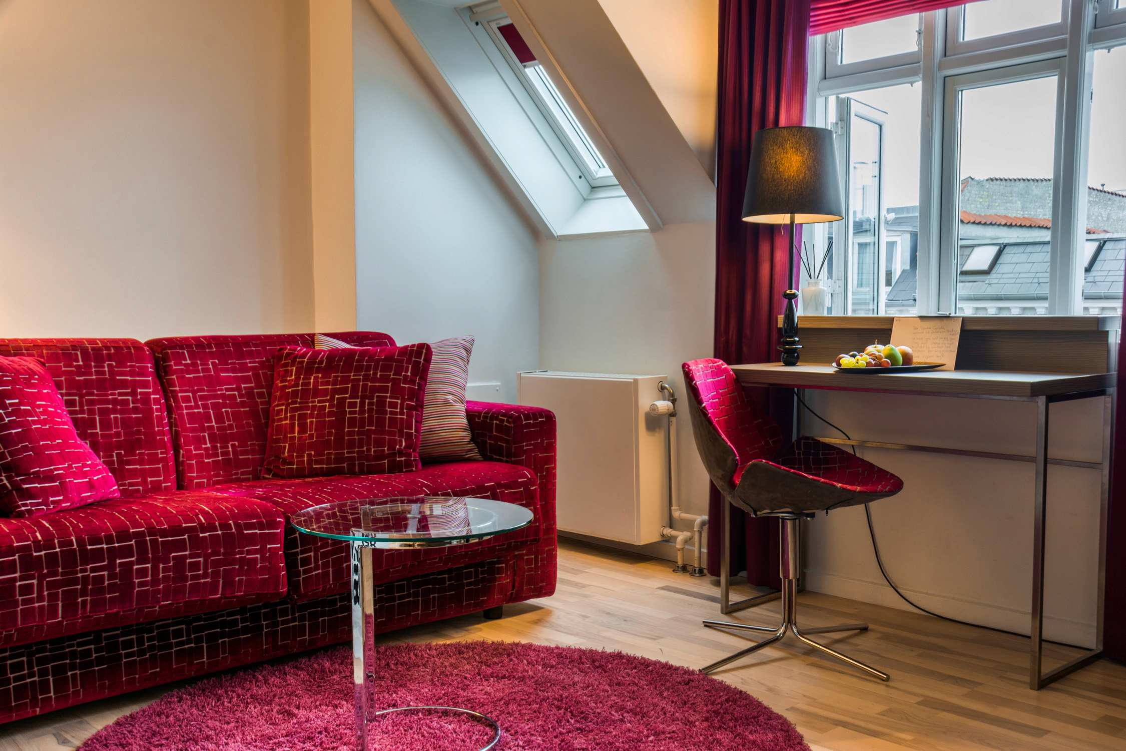 A modern hotel room features a red patterned sofa, a small round table, and a desk with a lamp near a large window.