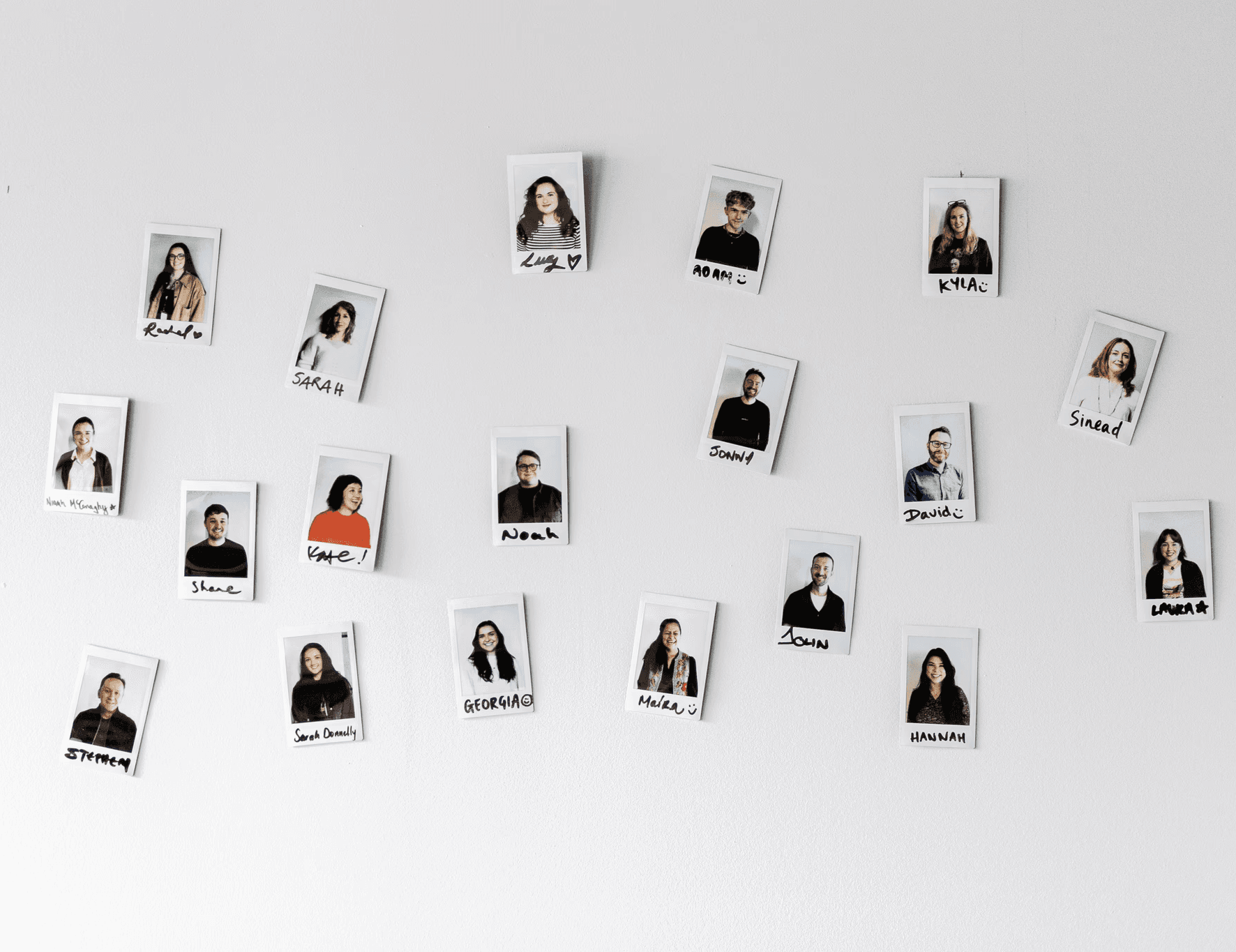 Polaroid-style photos of team members arranged on a white wall, each image labelled with their name