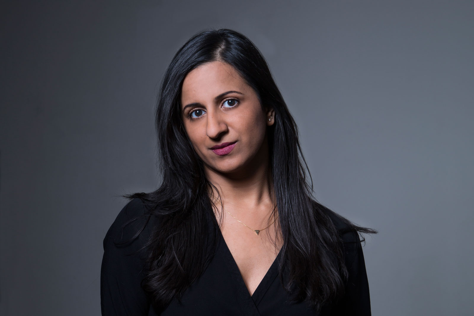 A woman with long, dark hair wears a black top and a delicate necklace with a small pendant. She looks directly at the camera with a neutral expression. The background is a solid, dark gray color.
