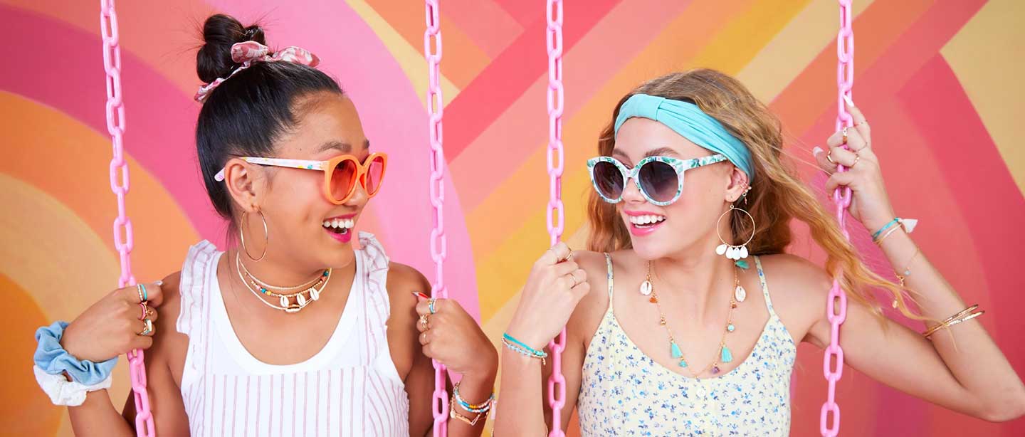 Two girls in sunglasses and layered jewelry laughing on swings against a pink and orange mural