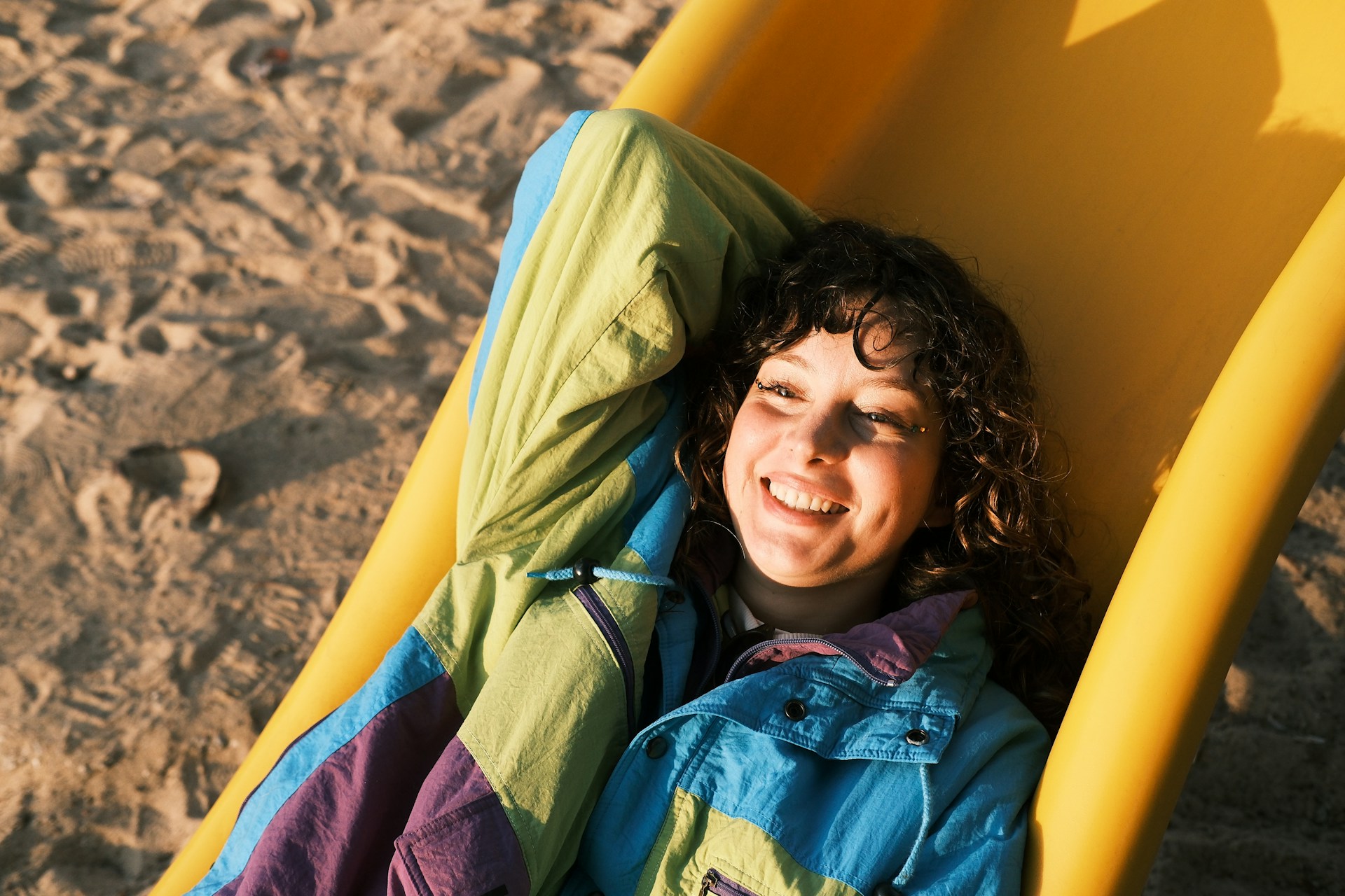 A high-angle shot of the woman lying down on a yellow slide at a beach, smiling broadly. She is wearing a colorful windbreaker, and the background shows footprints in the sand.