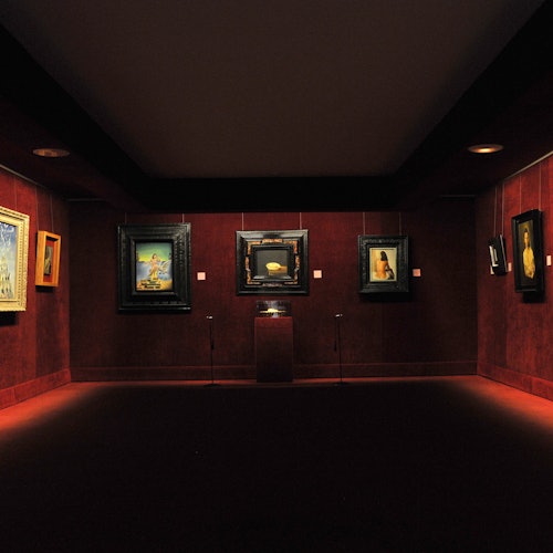 גלריית אמנות עם תאורה מעומעמת וציורים ממוסגרים על קירות אדומים, כאשר כן תצוגה עומד במרכז תחת אור הממוקד.