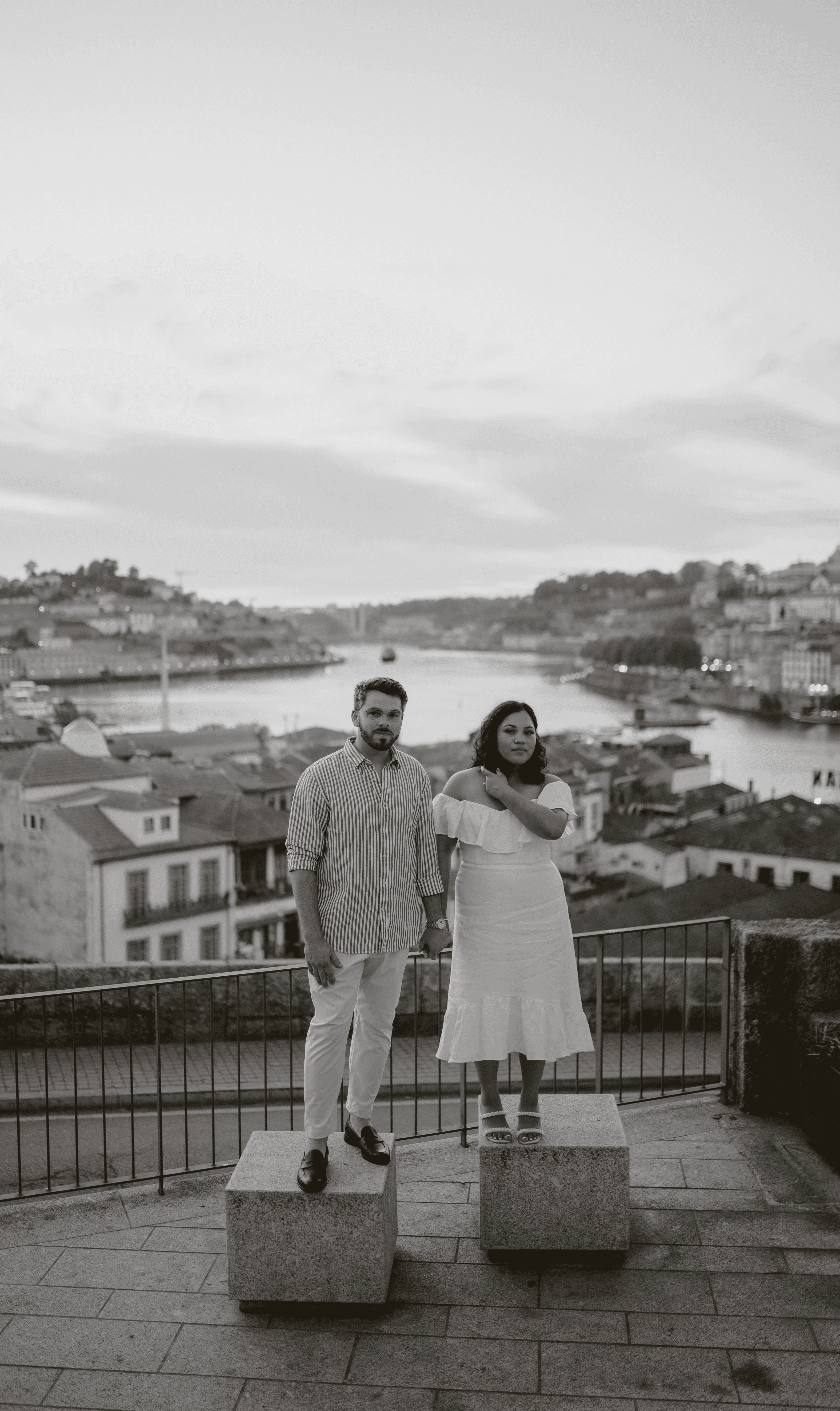 Black and white photo of a man and woman standing on stone blocks overlooking a cityscape and river. The man, dressed in a striped button-down shirt and light-colored pants, stands on the left. The woman, in an off-the-shoulder white dress, stands on the right with one hand on her shoulder. Behind them, a scenic view of a European city with buildings on hills and a river winding through the center is visible at sunset or twilight. A railing lines the overlook where they are standing.