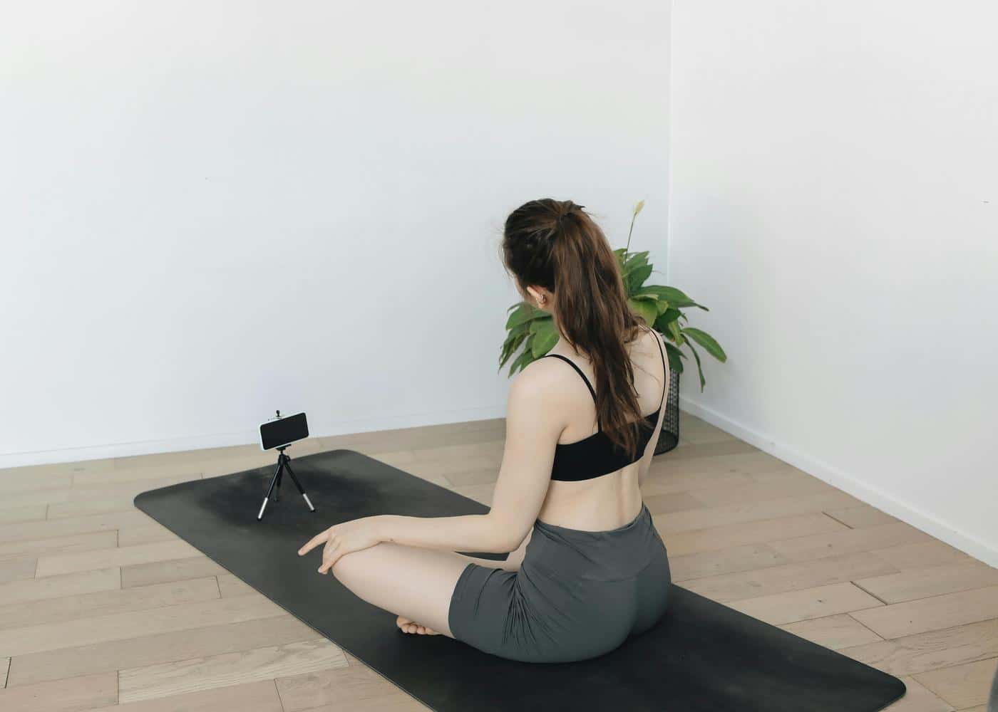 Woman sitting on black yoga mat looking at phone on tripod in front of her