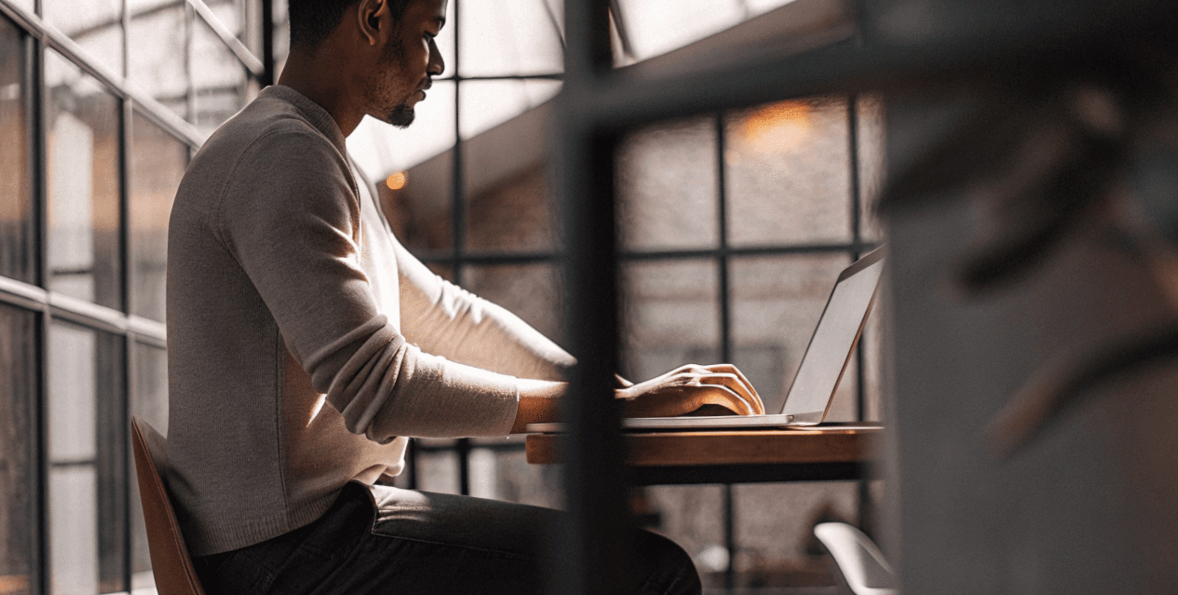 Man working on laptop near window. He is sitting at a wooden table in a cafe.