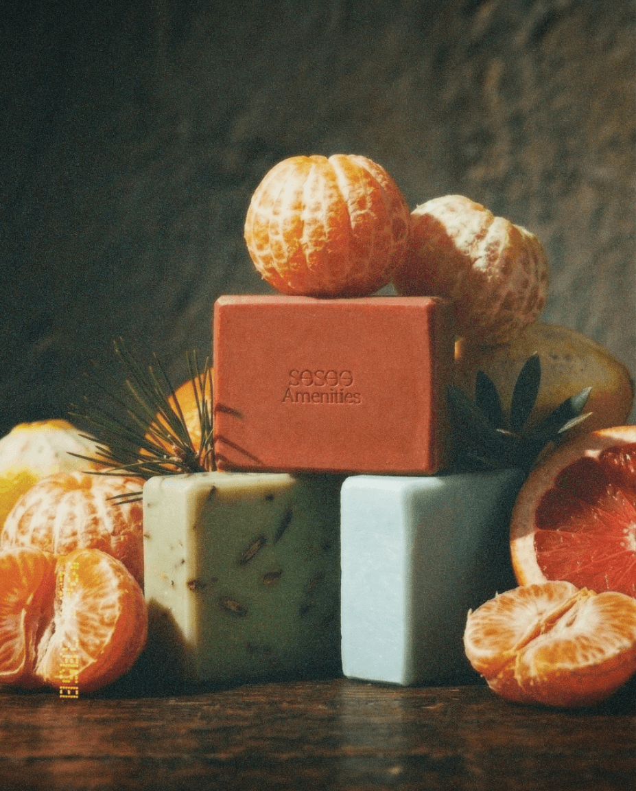 Assorted soaps in red, green, and blue are stacked with fresh citrus fruits, including tangerines and grapefruit, on a rustic wooden surface.