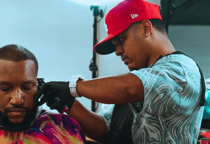 A barber shapes a client’s haircut using clippers near a bright ring light in barbershop. 