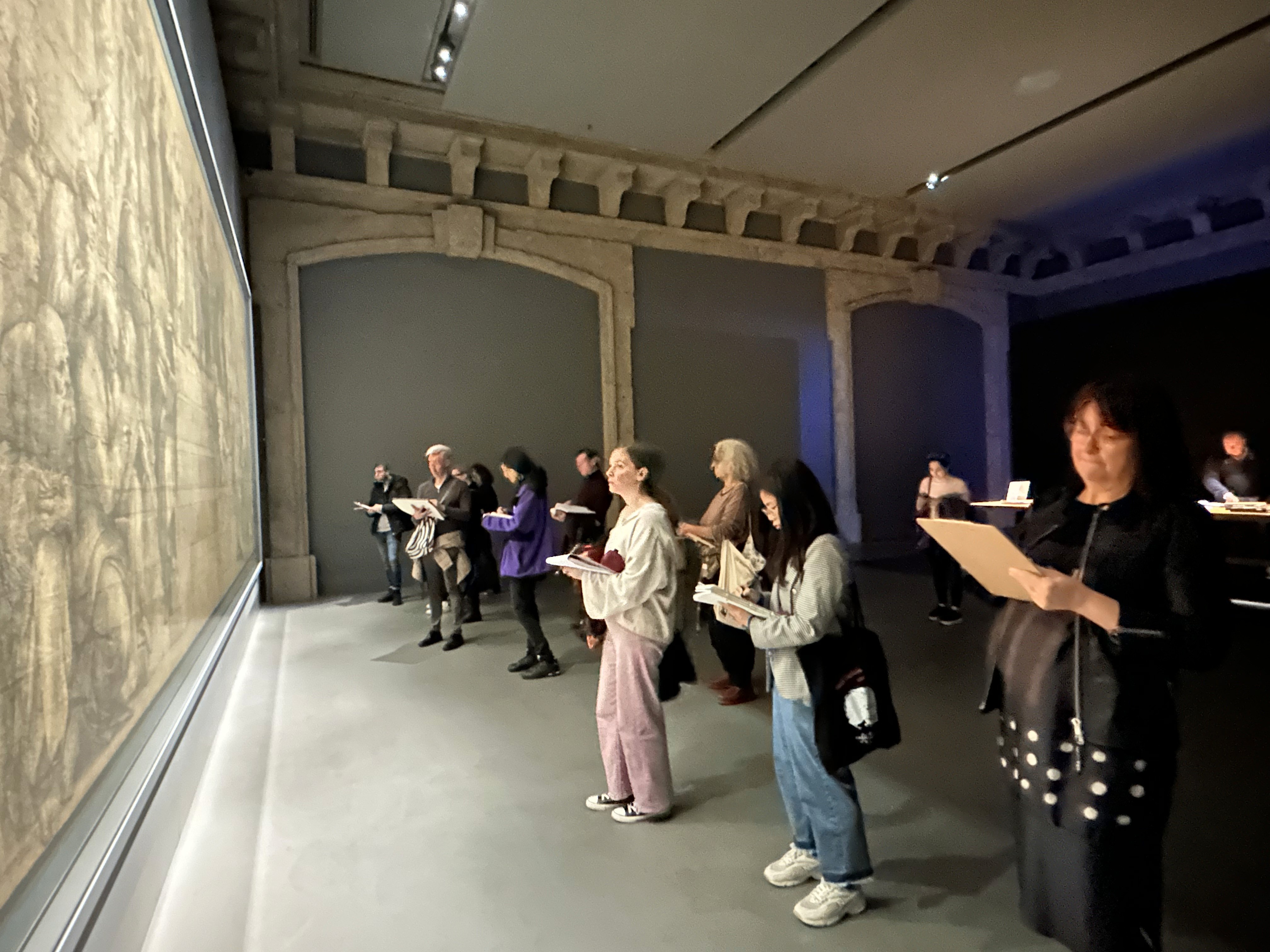 A group of people stand intently observing a large, detailed drawing at an art gallery, each holding sketchbooks and concentrating on replicating the artwork in the dimly lit, spacious room.