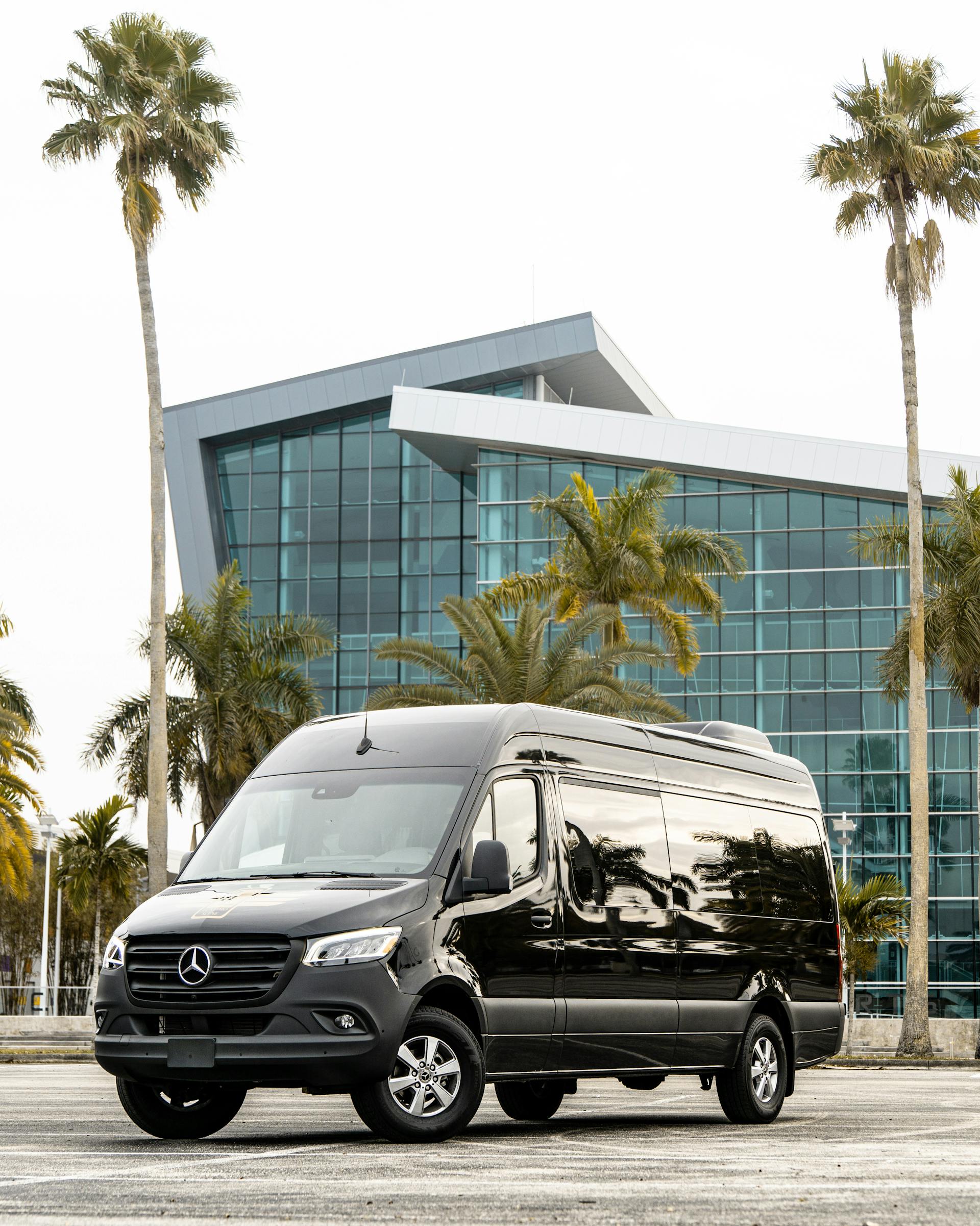 Luxury black Mercedes-Benz Sprinter van parked in front of a modern glass building, surrounded by palm trees, symbolizing premium transportation services for NYC tours.