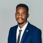 A person in a gray double-breasted suit and black turtleneck stands in warm lighting, facing forward. The expression is serious and composed.