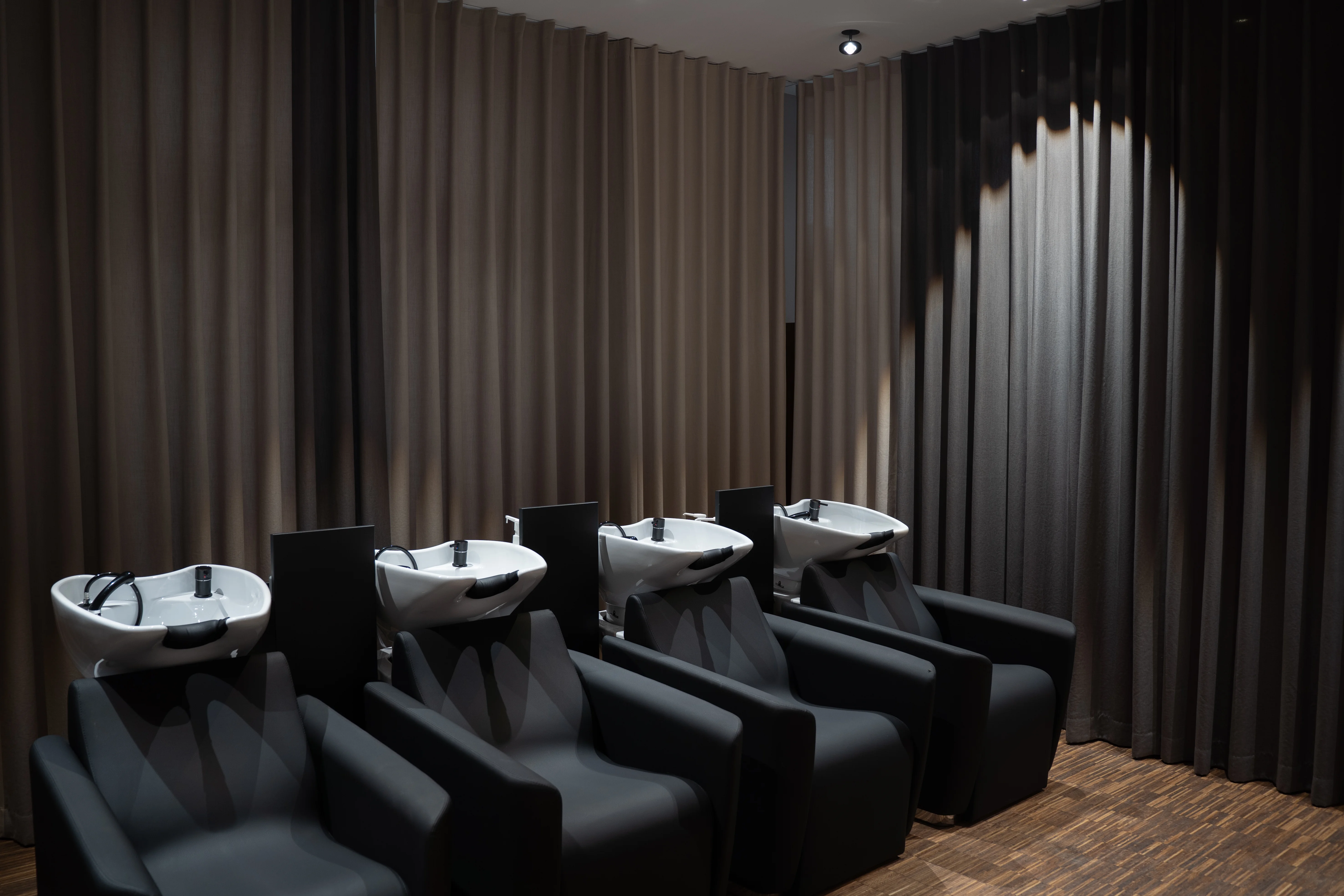 Hair-washing area at EMPL FRISEURE with four black reclining chairs and white basins, surrounded by floor-length curtains in beige and anthracite.